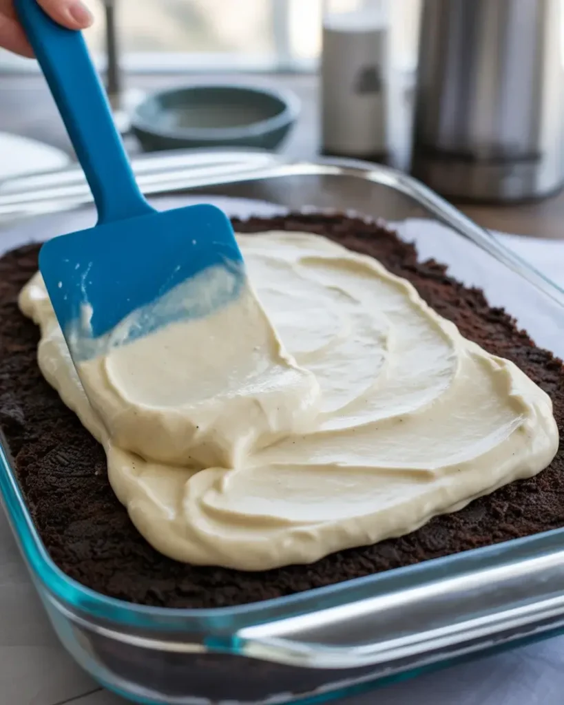 Spreading creamy filling over the pressed Oreo base in a 9x13 casserole dish